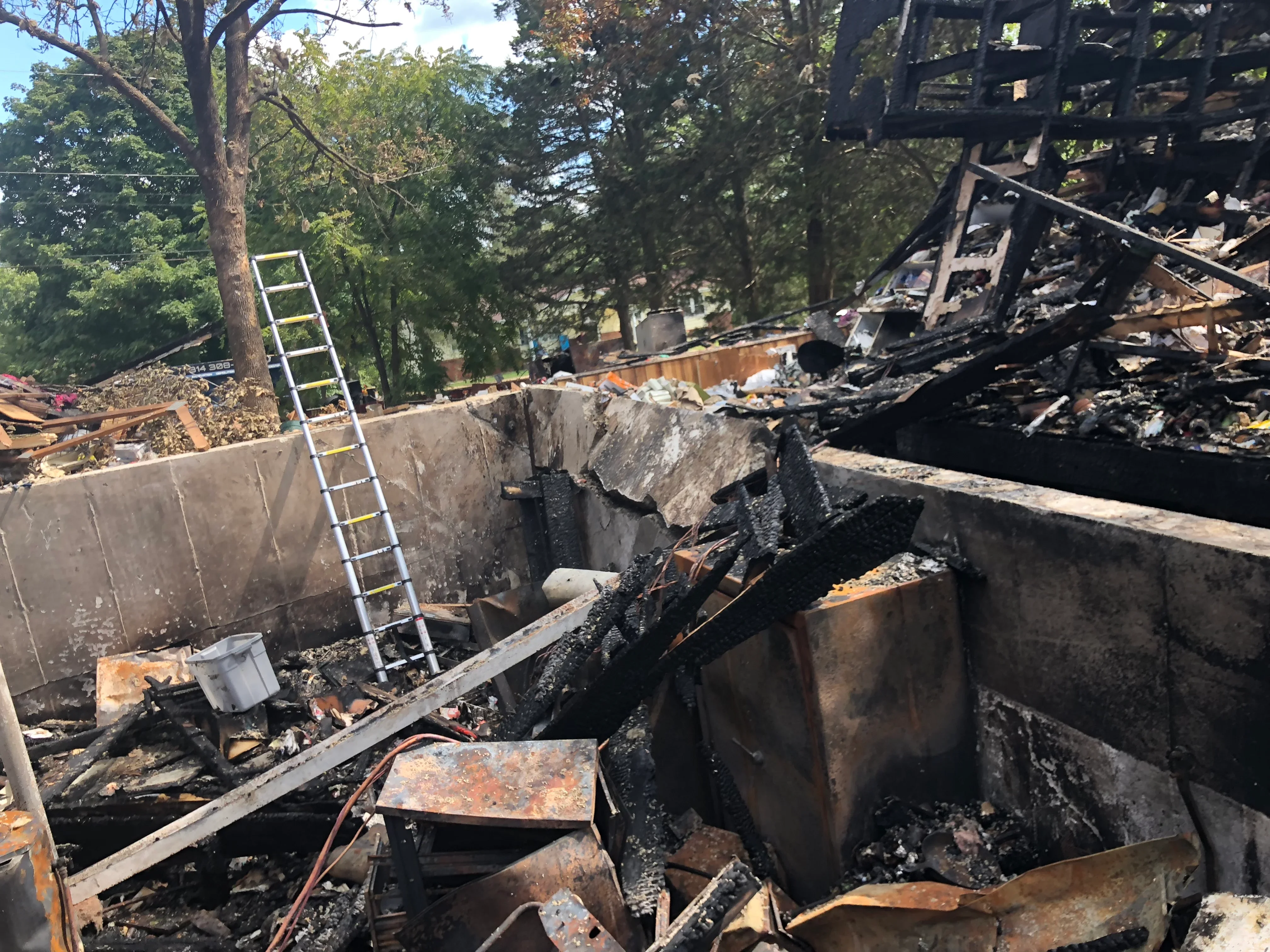 Demolition site with charred wood debris, concrete foundation, metal framing, and aluminum ladder