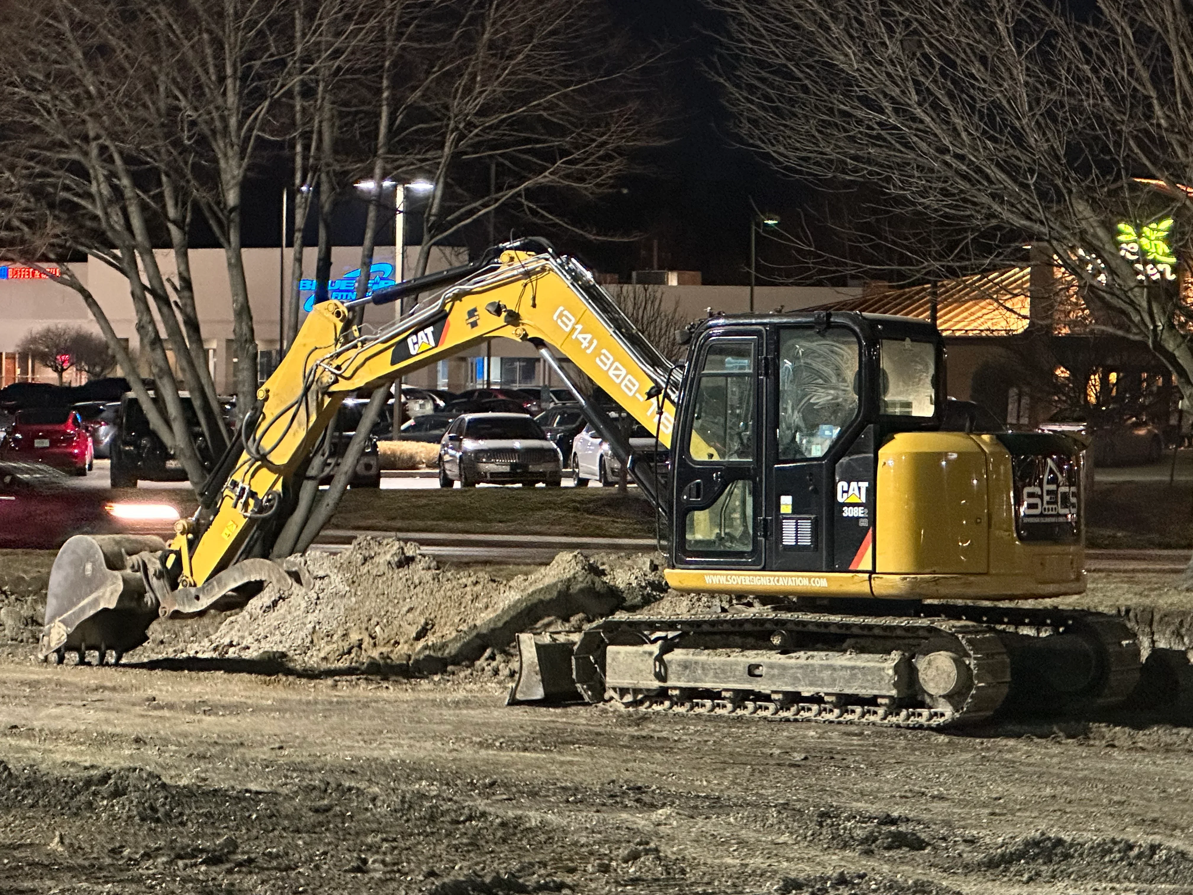 Yellow CAT 306E mini excavator with bucket, moving soil on commercial lot at night