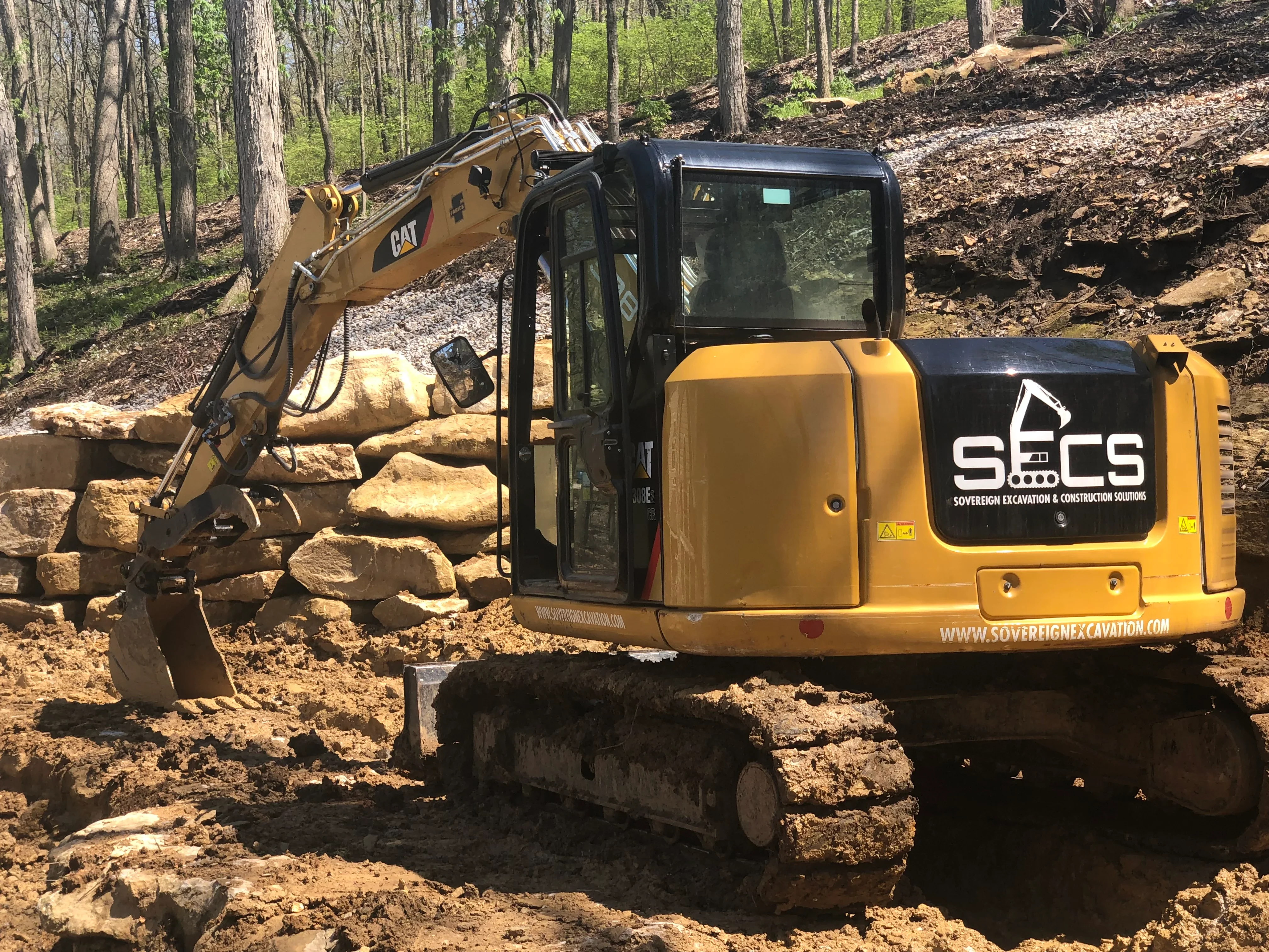CAT excavator with bucket removing logs and grading wooded site among standing trees